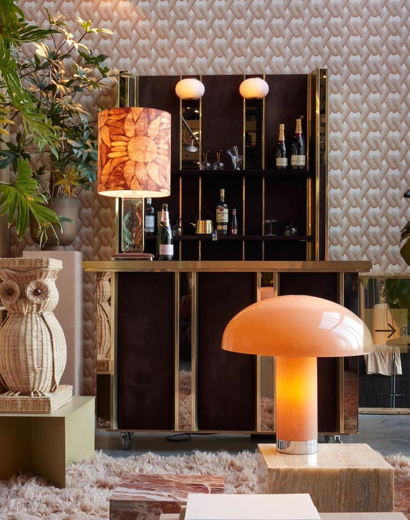 3, tall, colourful lamp bases in orange, silver, and green, all with a contrasting or matching lamp shade stood apon a shelving unit infront of a blue tilled wall