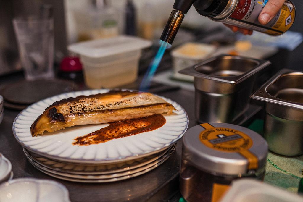 someone blow torching a topping on some toast apon a beautiful serax, scalloped edged, plate surrounded by little tins and jars of ingredients