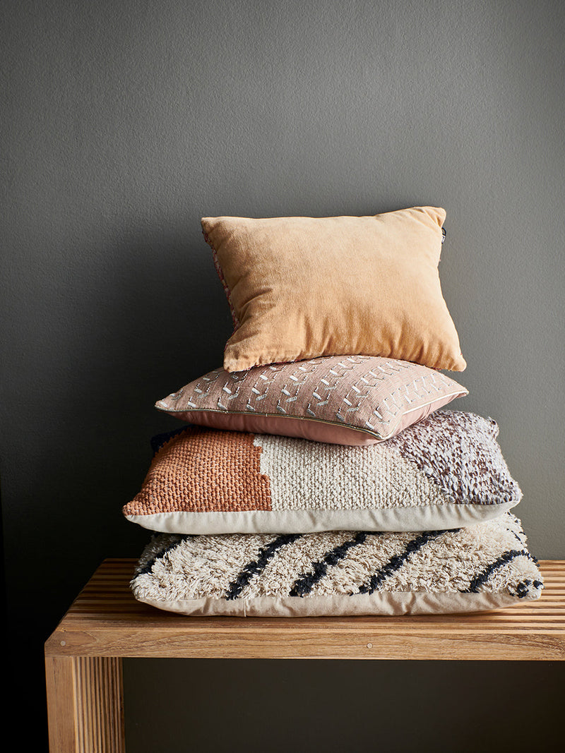 A stack of 4 cushions on a slatted bench made of a light wood. The cushions are are HKliving with a light orange velvety number, as well as a couple of fluffier options