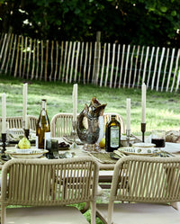 outdoor dining table set for a garden meal, with cream taper candles in dark metal candlesticks, patterned ceramic plates, lemons and wildflowers in a stoneware jug, and two chickens standing on the table