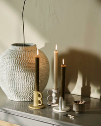 three lit taper candles in different ceramic candle holders beside a large textured stoneware vase, an olive green taper in a holder shaped like a letter B, a cream taper in a patterned holder, and an olive green taper in an arched holder, with a small bowl of matches alongside