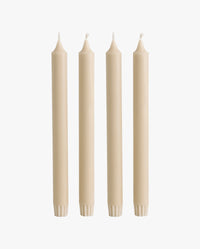 row of lit candles on a white mantelpiece shelf: two ceramic fish-shaped candlesticks holding cream taper candles, a cluster of cream and dark olive green pillar candles of varying heights, and a brass candlestick holding another taper candle, with framed artwork on the wall behind
