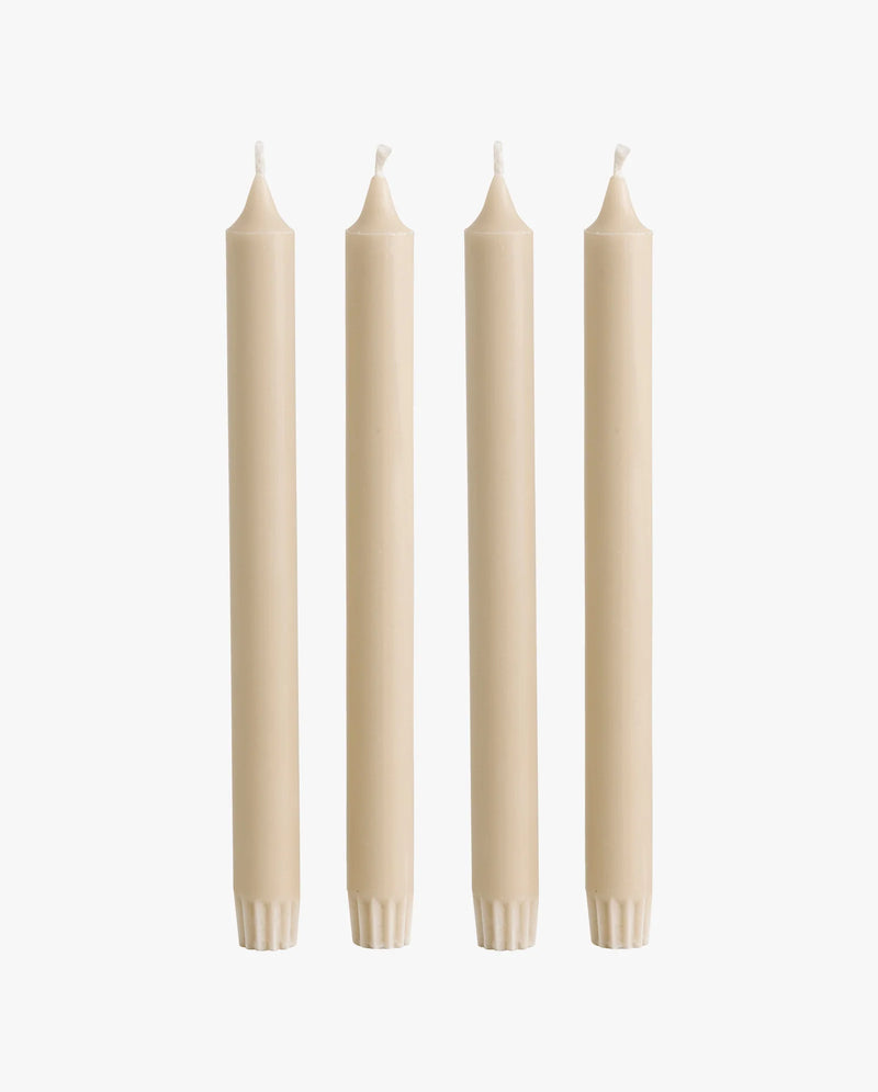 row of lit candles on a white mantelpiece shelf: two ceramic fish-shaped candlesticks holding cream taper candles, a cluster of cream and dark olive green pillar candles of varying heights, and a brass candlestick holding another taper candle, with framed artwork on the wall behind