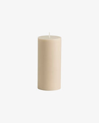 cluster of lit Nordal pillar candles in cream and dark olive green on a round marble table beside a cream bouclé chair, with a brass candlestick and a small ceramic dish among them