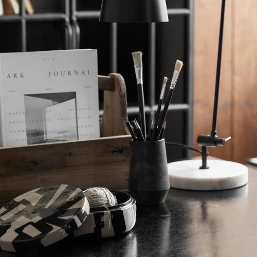 the black and white lipsi box being used on an office desk holding a ball of twine. A tall black pot sits behind it with paint brushes and an art book is open on the side