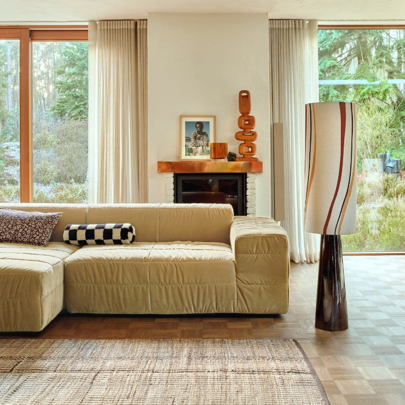 living room with a large squishy yellow sofa, big windows dressed with light curtains on the side, a fire and mantlepiece with a picture of alady in a stripy jumper and a tall wooden sculpture. The tall floor lamp that is styled similar to a table lamp with a wide bases and long tubular lamp shade with striped that curve gently down it in red, yellow, and black