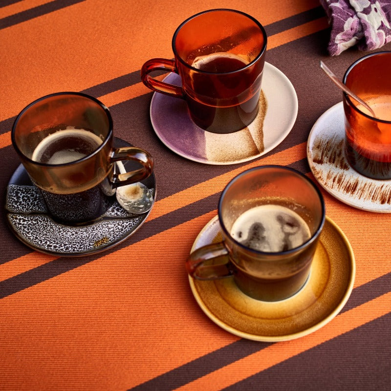 the four saucers being used with a set of handled, brown glass, mugs all with half drunk americano sat on a ornage and purple striped table cloth