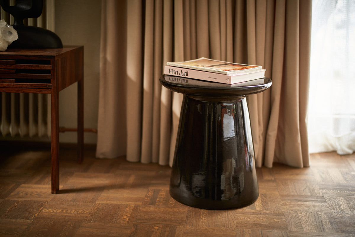 Glossy black HKLIVING earthenware side table with books on top beside a wooden chest of drawers