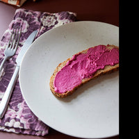 image of burgundy purple cloth napkin on a table with cutlery and a sandy plate also from hk with toast spread with a bright purple beetroot hummous