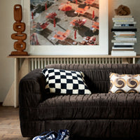 living room set up with a blakc soft sofa and a wooden artwork on a mantlepiece in the background. the sofa has 2 cushions on it including one that looks soft and fluffy in a checker board style in hk living style