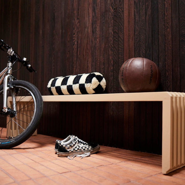 the white and black checkered bolster cushion on a slatted bench wiht a heavy brown leather medicine ball, checkered muddy trainers, and a mointain bike leaning: all with a back drop of a dark wood panel wall