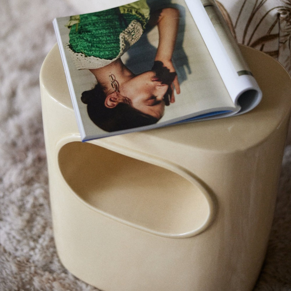 glossy cream side table with internal shelf which can be used of a stool tool from hkliving on a fluffy rug and with an open magazine on top revealing the image of a lady leaninig against a wall