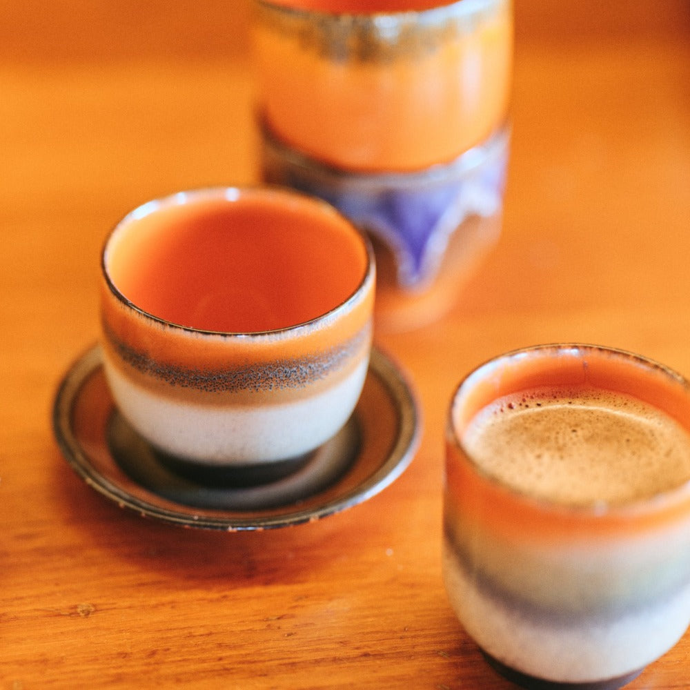 tea tme set-up with a stack of 70s colourful hkliving coffee cups, one on a saucer, and a different shaped mug with coffee in it. Lots of orange, purple, and browns