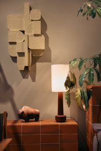 HKLIVING natural linen table lamp lit and glowing, styled on a terracotta tiled plinth beneath a wall sculpture and plant