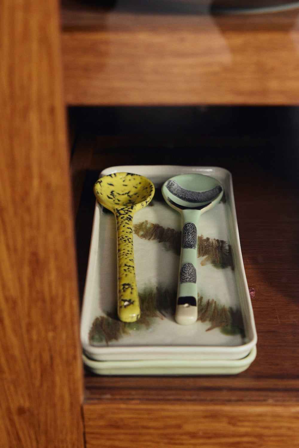 Two HKLIVING Sketch spoons resting on the Tide cake tray in green stoneware, displayed on a wooden cabinet shelf