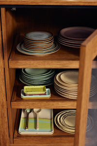 Open wooden cabinet shelving displaying stacked HKLIVING 70s Ceramics plates and trinket dishes in multiple colourways