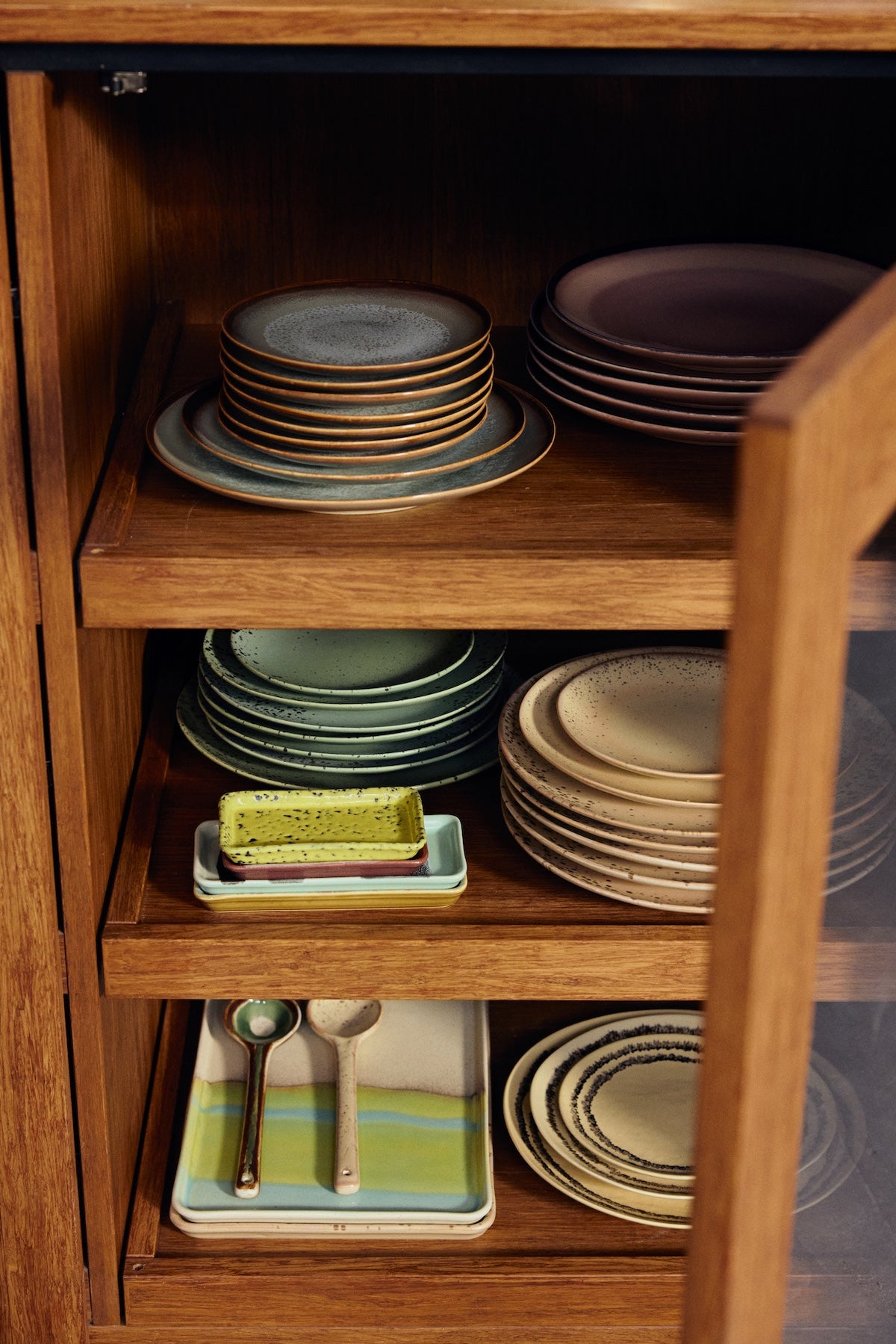 Open wooden cabinet shelving displaying stacked HKLIVING 70s Ceramics plates and trinket dishes in multiple colourways