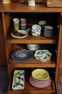 The HKLIVING Tide cake tray with spoons displayed on the bottom shelf of an open wooden cabinet, alongside stacked bowls, plates and other 70s ceramics pieces