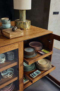 The medium HKLIVING teak cube displayed on a wooden sideboard alongside stacked ceramics and a table lamp, with an open cabinet of plates beneath