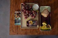 A speckled yellow HKLIVING Grit tray styled with a nut and dried fruit tray, a plate of red grapes and passionfruit, and a green tray with a sandwich, kiwi and passionfruit on a wooden table
