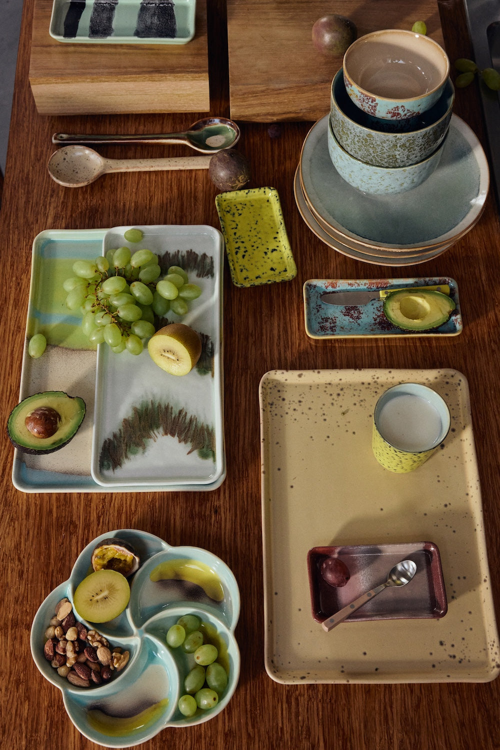 An overhead flat lay of the HKLIVING Grit collection on a wooden table, including the yellow large tray with a glass of milk, alongside speckled bowls, plates, spoons and other trays in the range