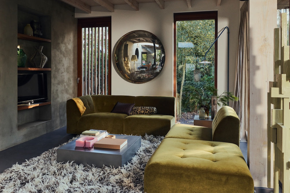 Green velvet corner sofa in a living room with the HKLIVING Panther bolster cushion tucked against a brown leather armchair in the background