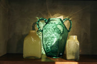 Green glass amphora hkliving vase styled on a shelf with smaller glass vessels, lit by sunlight