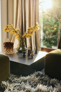 Olive green faceted glass Silhouette vase styled with yellow orchid stems on a dark coffee table, alongside smaller ceramic and glass vases in a sunlit living room