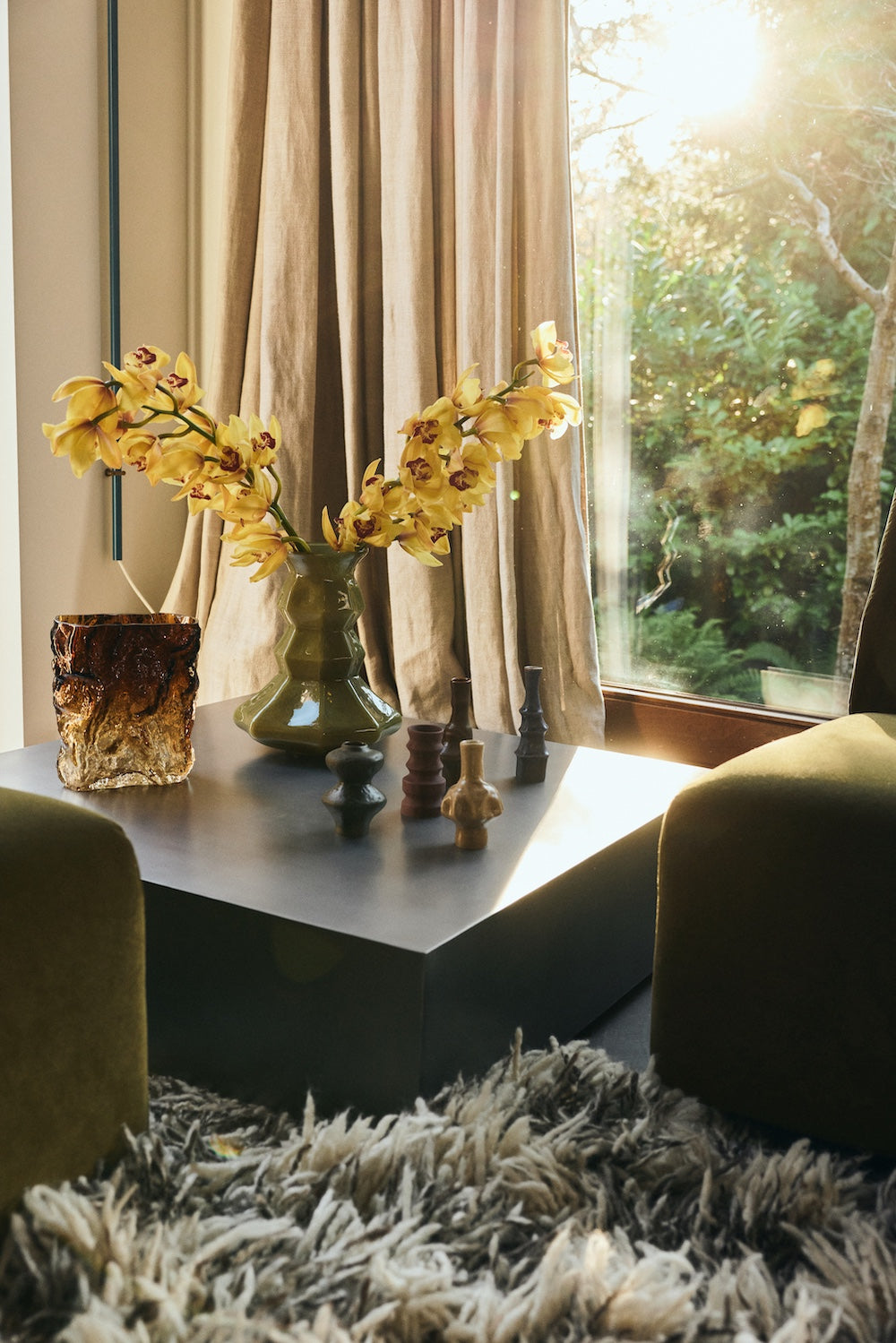 Olive green faceted glass Silhouette vase styled with yellow orchid stems on a dark coffee table, alongside smaller ceramic and glass vases in a sunlit living room