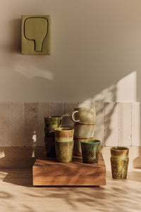 A stack of speckled stoneware cups and mugs displayed on the large HKLIVING teak cube, styled beneath a wall-mounted ceramic artwork