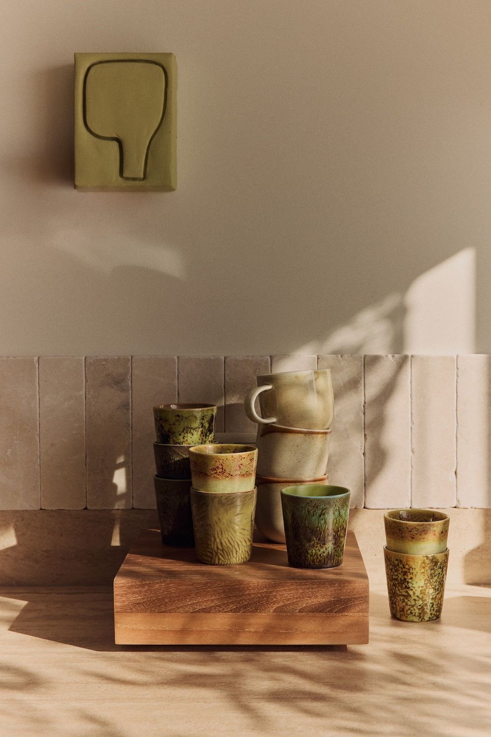 A stack of speckled stoneware cups and mugs displayed on the large HKLIVING teak cube, styled beneath a wall-mounted ceramic artwork