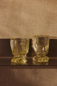 Two HKLIVING Citrine glasses displayed on a dark shelf against a woven beige fabric backdrop, showing their faceted glass form