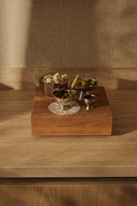 Silver bowls of olives, gherkins and popcorn arranged on top of the large HKLIVING teak cube, styled against a woven curtain backdrop