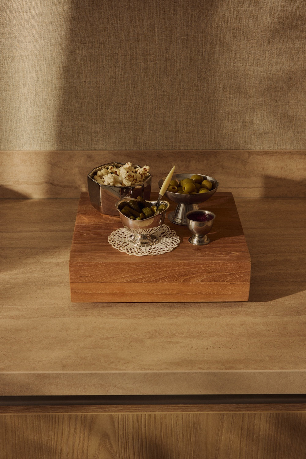 Silver bowls of olives, gherkins and popcorn arranged on top of the large HKLIVING teak cube, styled against a woven curtain backdrop