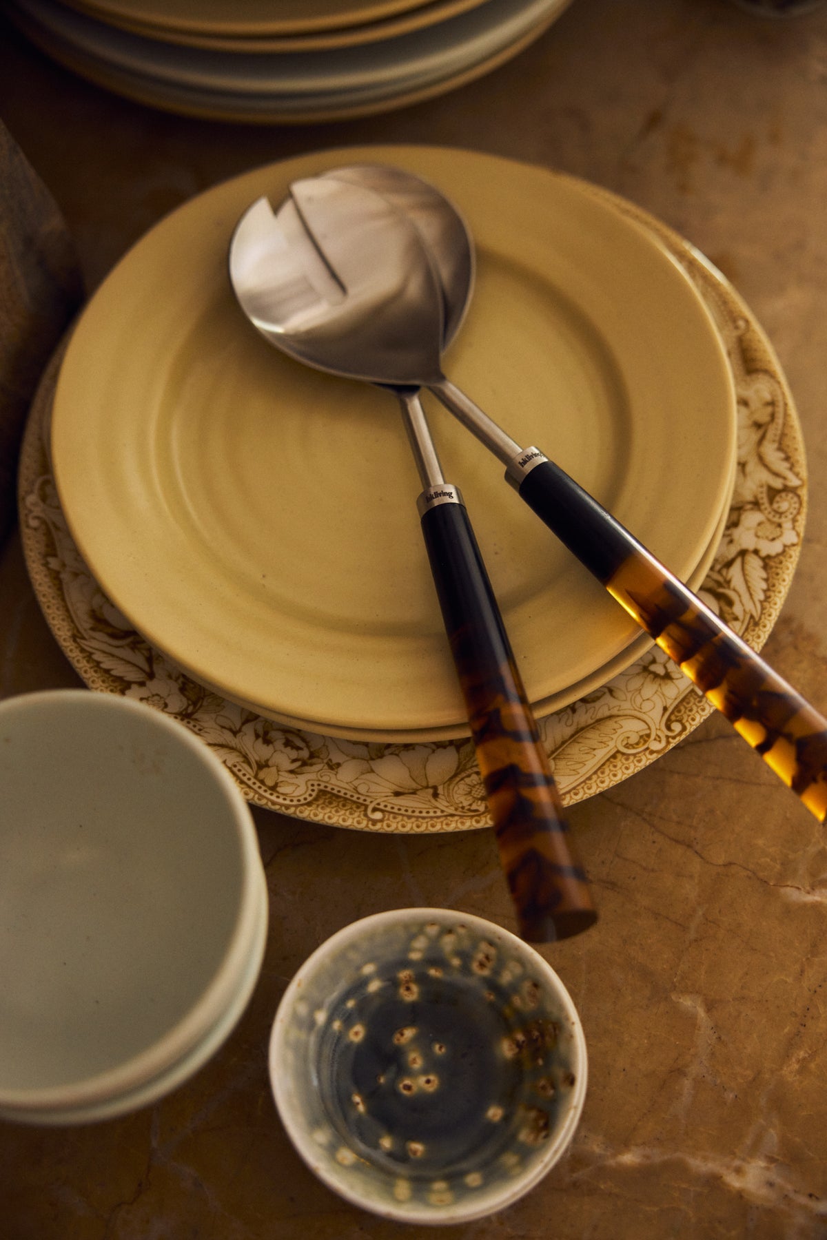 Havana salad servers resting across a stack of mustard-yellow and patterned plates, styled with ceramic bowls and a small glazed dish