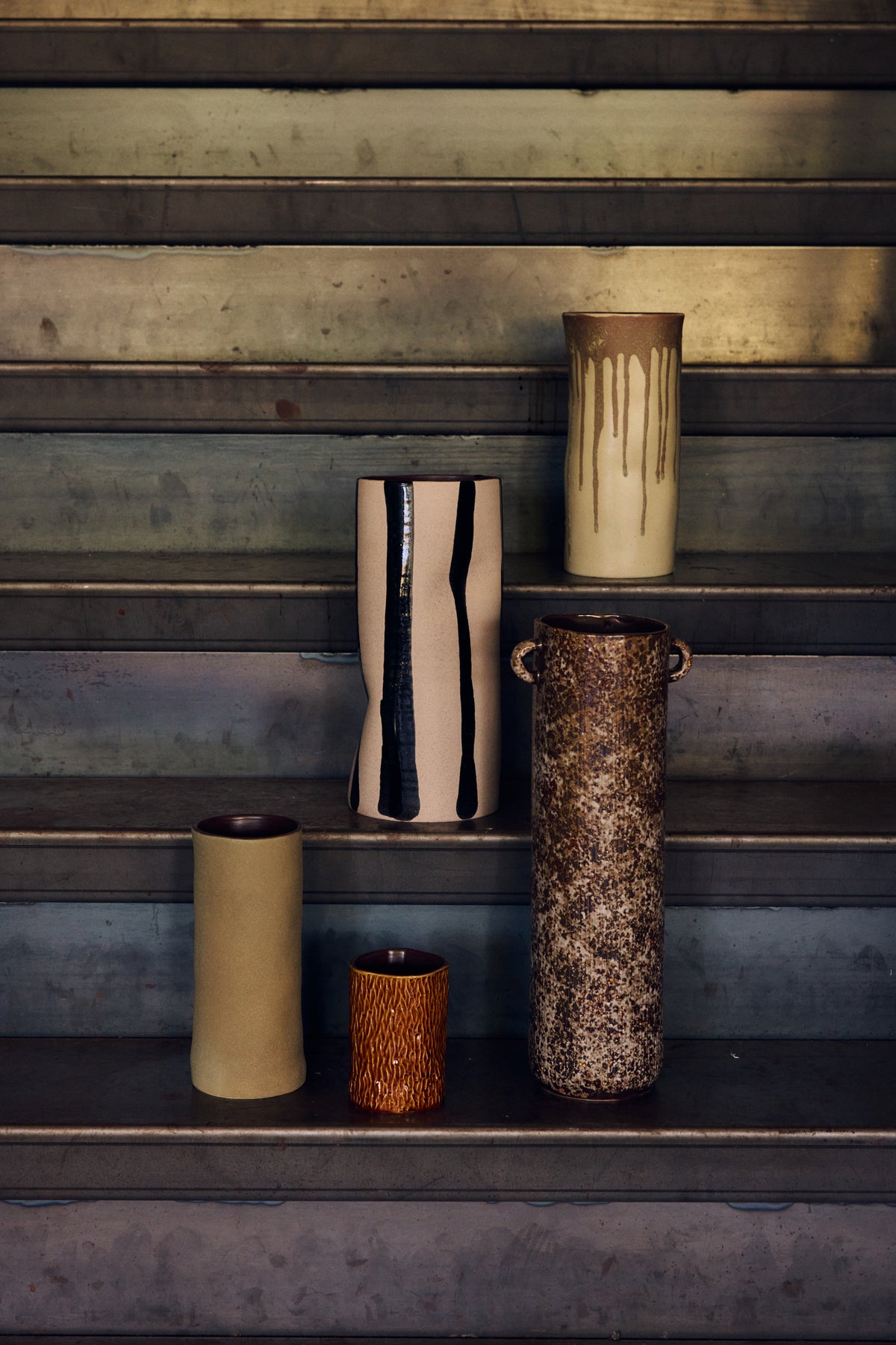 The Long Vase, Reactive Brown by HKLIVING displayed on metal steps alongside other tall ceramic vases in cream, pink and olive tones