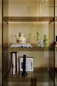 The Mono Vase, Sand/Black by HKLIVING styled on open glass shelving with a black ribbed candlestick, next to a yellow ceramic pitcher and green glass candlesticks above