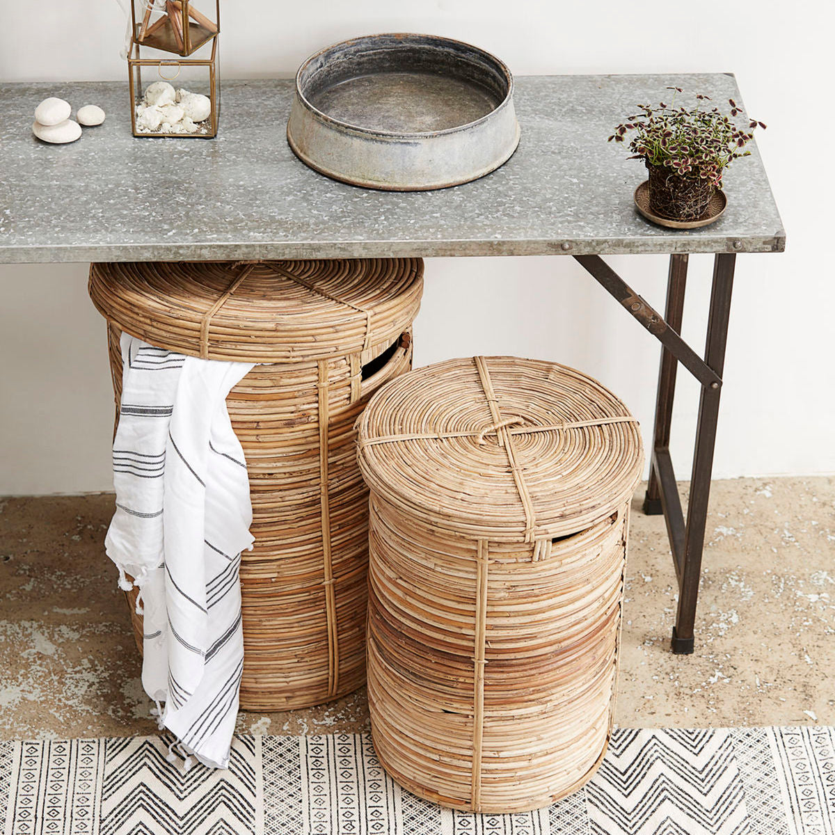 Two House Doctor Chaka woven laundry baskets with lids styled under a console table in a bathroom, with a striped towel draped over one basket