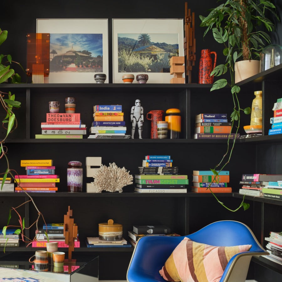 a colourful snug surrunded with shelves which are black to match the wall behind but the cooour from the ceramics, decorative pieces, and the electric blue armchair really bring the image to life