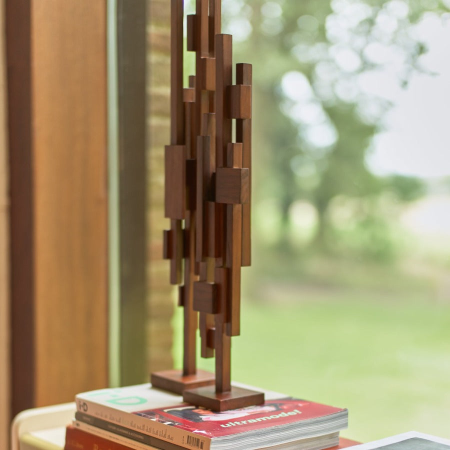 2 of the tall teak wood scultpures next to each other stood on a small stack of books on a book shelf in a large window overlooking a woodland in summer