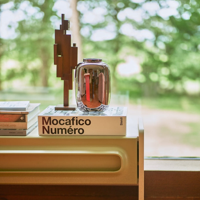 small scultpure looking almost robotic next to a shiny metalic vase on a large book