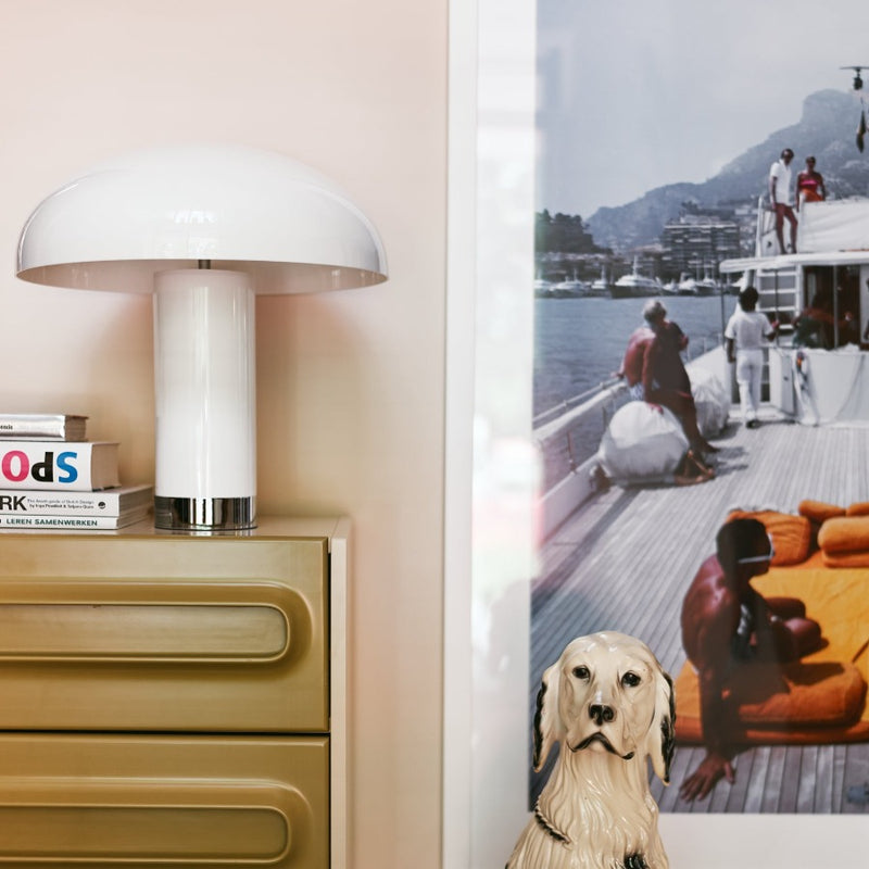 a pure white mushroom lamp stands on a coffee coloured chest of drawers form hk living with books and a paiting on the wall of people relaxing on the deck of a boat creating a joyfull ambiance with a personal style