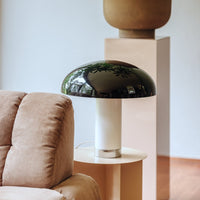 muchroom lamp stands on a side table next to a light brown soft sofa. with its black rounded lamp shade and white base which is fnished at the bottom with a chrome colar all reflecting the scene aaround it including other hk living items