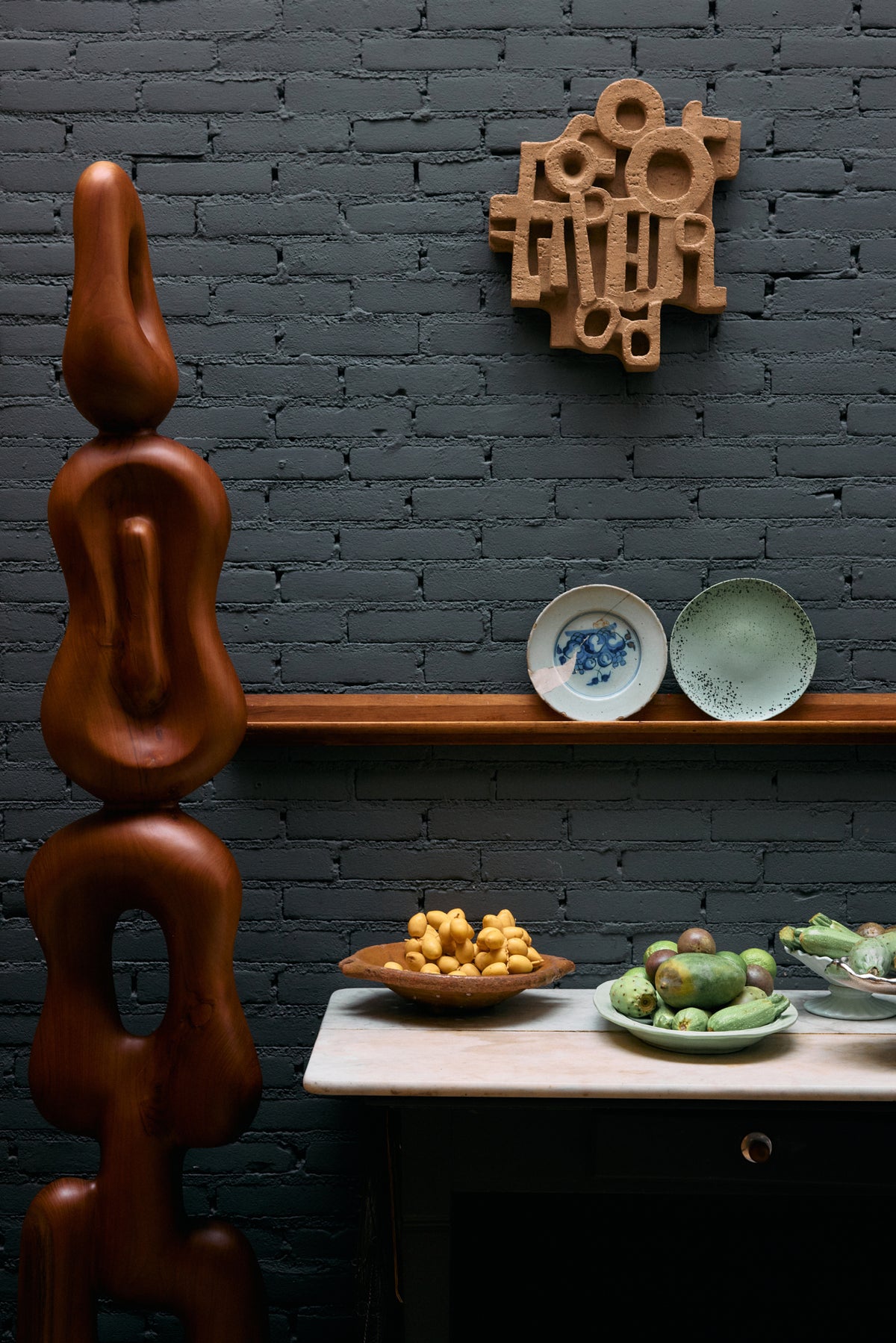 HKLIVING brutalist terracotta wall sculpture on a dark brick wall above a shelf of plates and a sculptural wooden figure