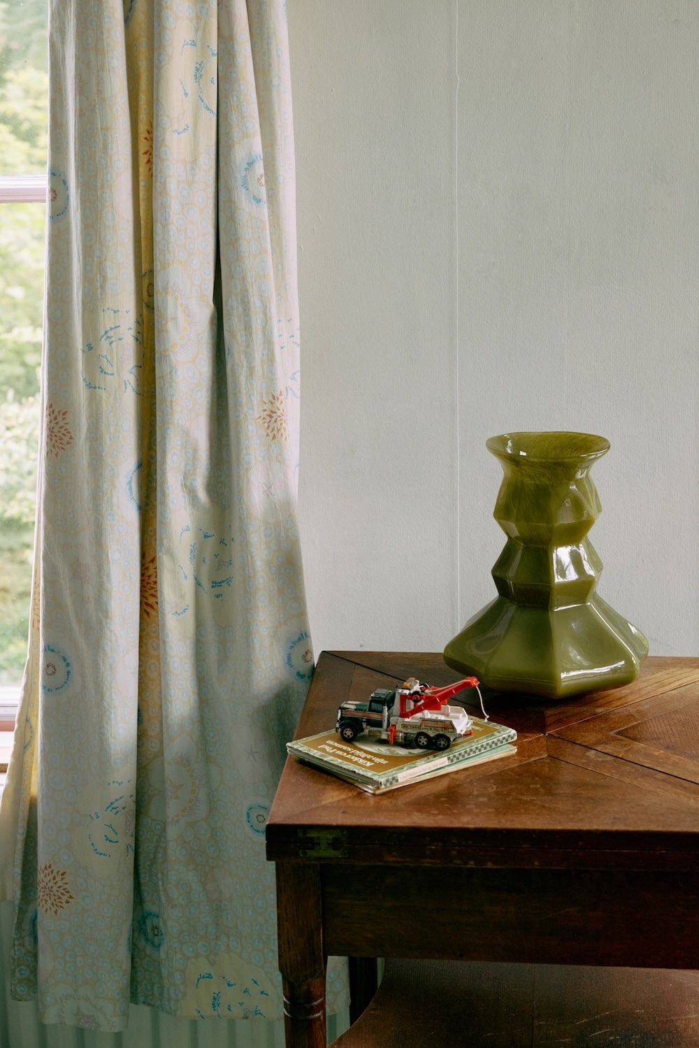 Olive green Silhouette glass vase displayed empty on a wooden side table beside a patterned curtain and a vintage toy truck