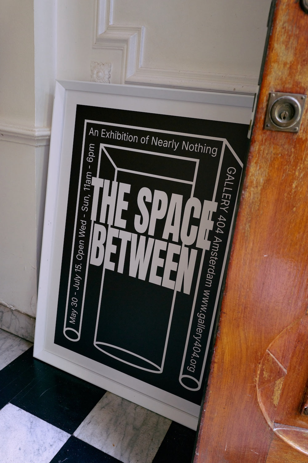 Between Space framed poster by HKLIVING propped against a door in a black and white tiled hallway