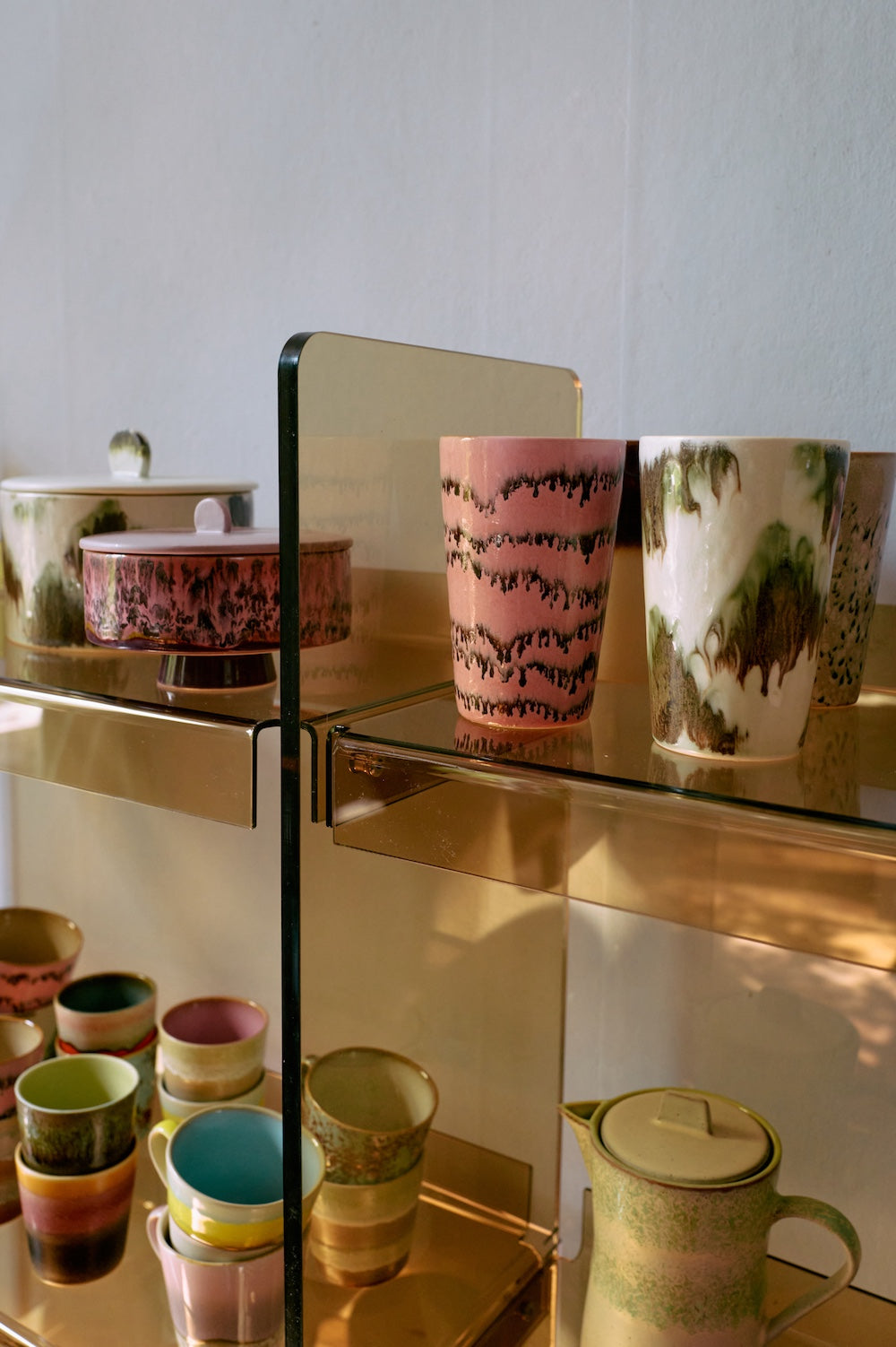 HKLIVING 70s ceramics pieces displayed on a glass and brass shelving unit, including the Tide tea mug alongside a pink mug and mixed coloured cups below