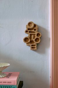 The HKLIVING brutalist wall sculpture hung on a pale pink plaster wall above a stack of books and a scalloped ceramic dish