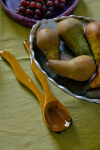 The HKLIVING ochre salad cutlery resting beside a bowl of pears on a green linen tablecloth, with a bowl of red grapes in the background