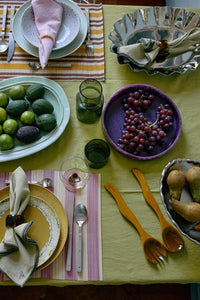 The HKLIVING ochre salad cutlery styled as part of a full table setting on a green linen tablecloth, with grapes, avocados, limes and patterned place settings